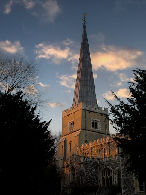 The image features the spire of Saint Mary Church Harefield rising sharply into the sky, with the church's stone and brick facade visible at the base. The scene is set during the day with a partly clo
