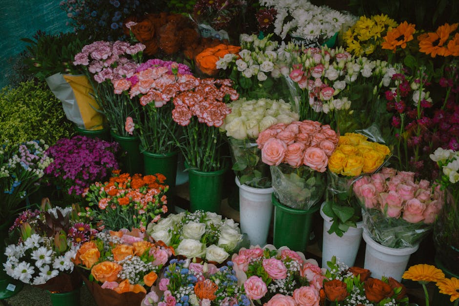 A vibrant display of various fresh flowers arranged in green, white, and transparent wrapping, including pink roses, yellow roses, white roses, orange carnations, purple asters, white daisies, and pin