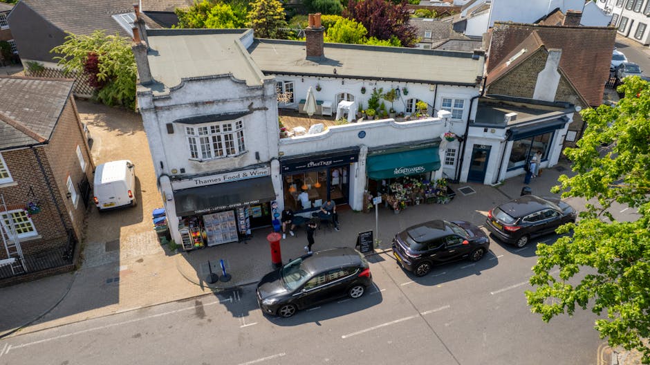 An aerial view of a charming row of small shops on Harefield High Street UB9, featuring a flower shop with a green and white awning, displaying colorful floral arrangements outside. Nearby, there is a