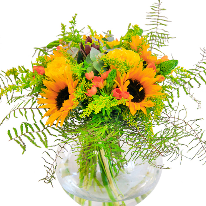 The spirit of Harefield's vibrant landscapes is elegantly gathered in this fiery floral arrangement, where grand sunflowers flare outwards, their bronze eyes reflecting the late afternoon sun. Yellow garden roses, each intricately layered, nestle close among the blooms, injecting the bouquet with classic charm and plush, spiraled texture. Throughout, the cheerful red orbs of Hypericum berries sparkle, creating moments of whimsy and depth, much like the wild berries found along the quiet trails of Harefield Moor. Encircling the bouquet, feathery fronds and dancing sprays of goldenrod lend movement and a breezy countryside feel. Hints of eucalyptus thread through the foliage with a flash of blue-green, adding fragrance reminiscent of cool, shaded copses off the Rickmansworth Road. Each flower and leaf is rooted in a crystal-clear, spherical glass vase whose watery embrace reveals the bouquet's vivid stems, amplifying the sensation of garden-fresh abundance. The interplay of textures-from suede sunflower petals to glossy berries and the crisp snap of greens-creates a tactile richness that delights both eye and imagination. Delicate, uplifting scents hint at open fields and tranquil moments near Harefield's canal. Thoughtfully composed for birthdays, anniversaries, or everyday joys, this arrangement is a testament to Harefield florists' expert touch and affection for their local community.